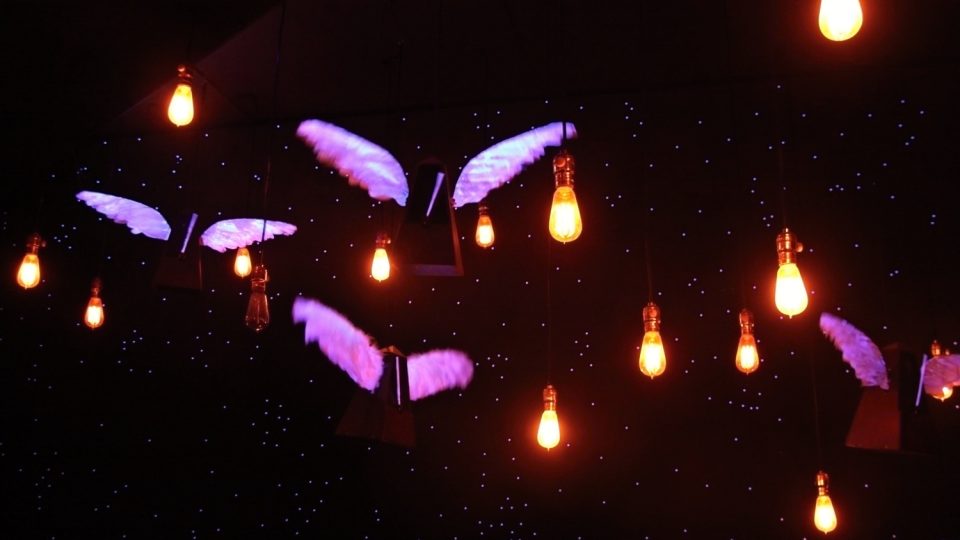 V potemnělém sále se vznášející létající metronomy se svítícími bílými křídly